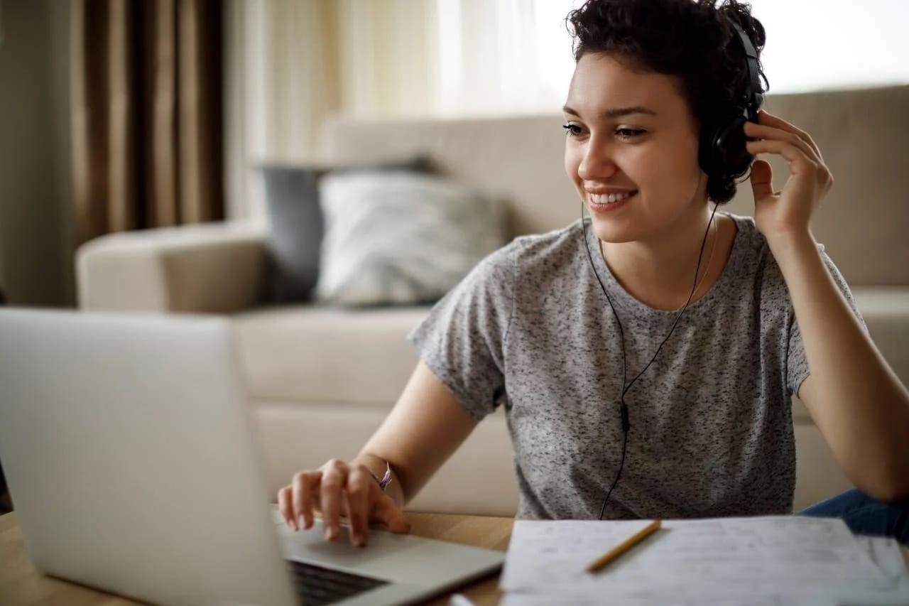 A woman with headphones sits at a table, focused on her laptop, immersed in her work or study.